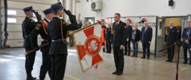 Nowy Komendant Powiatowy PSP w Grajewie stoi przed sztandarem komendy. Poczet salutuje. W tle zaproszeni goście. 