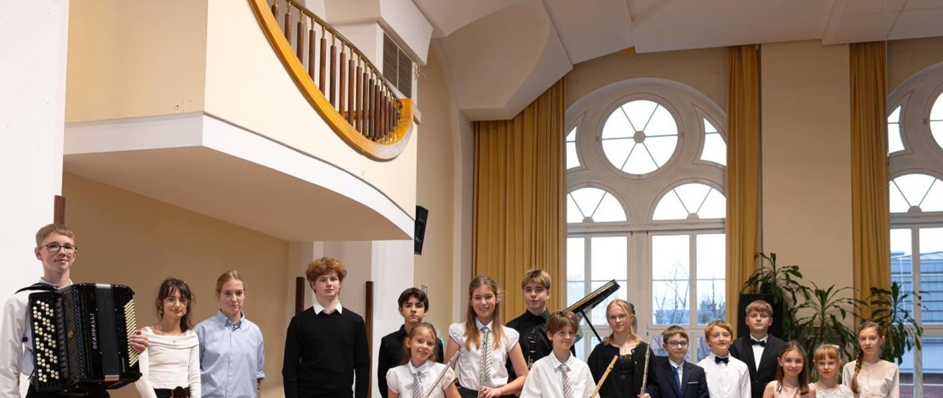 Zdjęcie kolorowe. Grupa kilkunastu osób stoi na drewnianym parkiecie w jasnej sali koncertowej. Po lewej stronie chłopak w okularach trzyma czarny akordeon marki Scandalli, a obok niego dziewczyna trzyma gitarę klasyczną. W centralnej części i po prawej stronie znajduje się grupa dzieci i młodzieży, z których wiele trzyma srebrne flety poprzeczne. Osoby są ubrane w stroje galowe: białe koszule, krawaty, ciemne spodnie, spódnice oraz wizytowe sukienki. W tle fragment fortepianu oraz wysokie okna z żółtymi zasłonami, przez które wpada dzienne światło. Nad grupą po lewej stronie widoczny jest biały balkon o zaokrąglonym kształcie z drewnianą balustradą. W lewym dolnym rogu umieszczono biały napis „aMuz”.