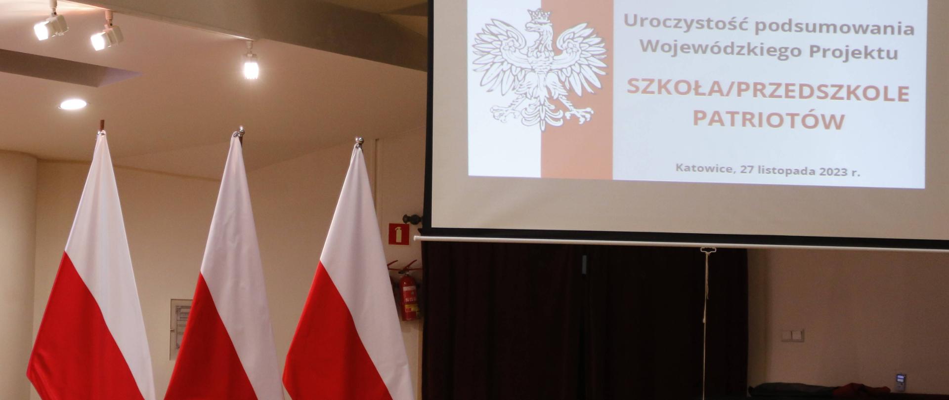 Po lewej widoczne trzy biało-czerwone-flagi, po prawej opuszczony ekran multimedialny z wyświetlonym slajdem, po którego lewej stronie znajduje się orzeł w koronie na tle biało-czerwonej flagi a po prawej tytuł uroczystości i data wydarzenia