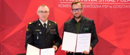 Dwóch mężczyzn pozuje do zdjęcia na tle czerwonego banera z logo Państwowej Straży Pożarnej. Jeden z nich ubrany jest w mundur strażacki (komendant wojewódzki PSP), drugi w ciemną marynarkę w kratę i zieloną koszulę (kurator oświaty). Obaj trzymają podpisane dokumenty porozumienia.