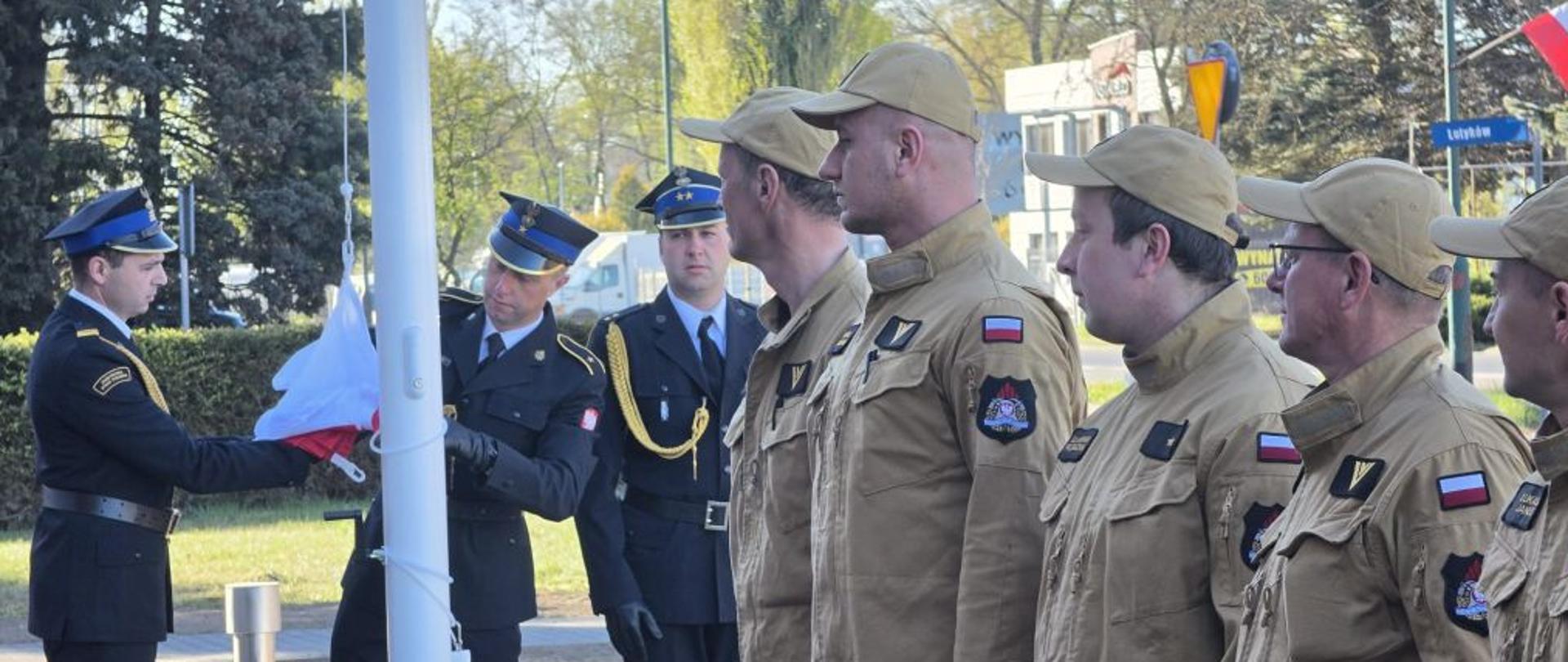 Zbliżenie na grupę strażaków podczas uroczystego wciągania flagi na maszt. Po lewej stronie dwaj strażacy w ciemnogranatowych mundurach wyjściowych przygotowują biało-czerwoną flagę. Po prawej stronie w równym szeregu stoją strażacy w piaskowych mundurach służbowych i czapkach z daszkiem, zachowując postawę zasadniczą. Na ich ramionach widoczne są naszywki z flagą Polski.