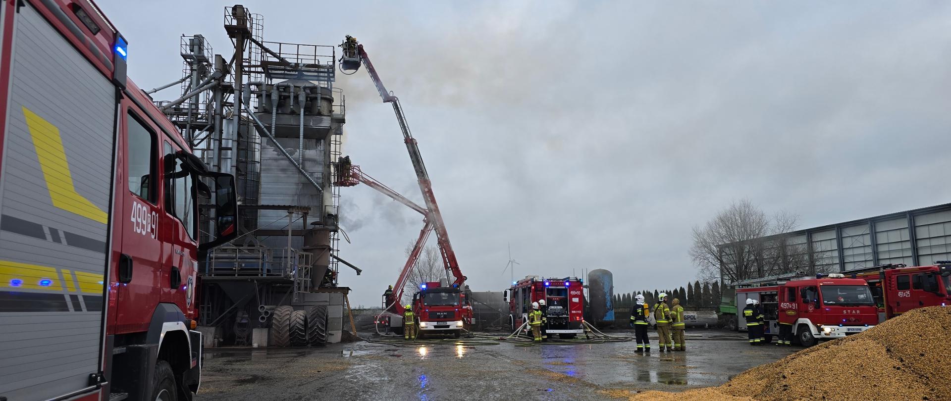 Pożar suszarni do zboża. Działania gaśnicze prowadzone z użyciem dwóch podnośników hydraulicznych, które sprawiono przy suszarni, w pobliżu auta gaśnicze. Na placu widoczna kukurydza usunięta z urządzenia. 