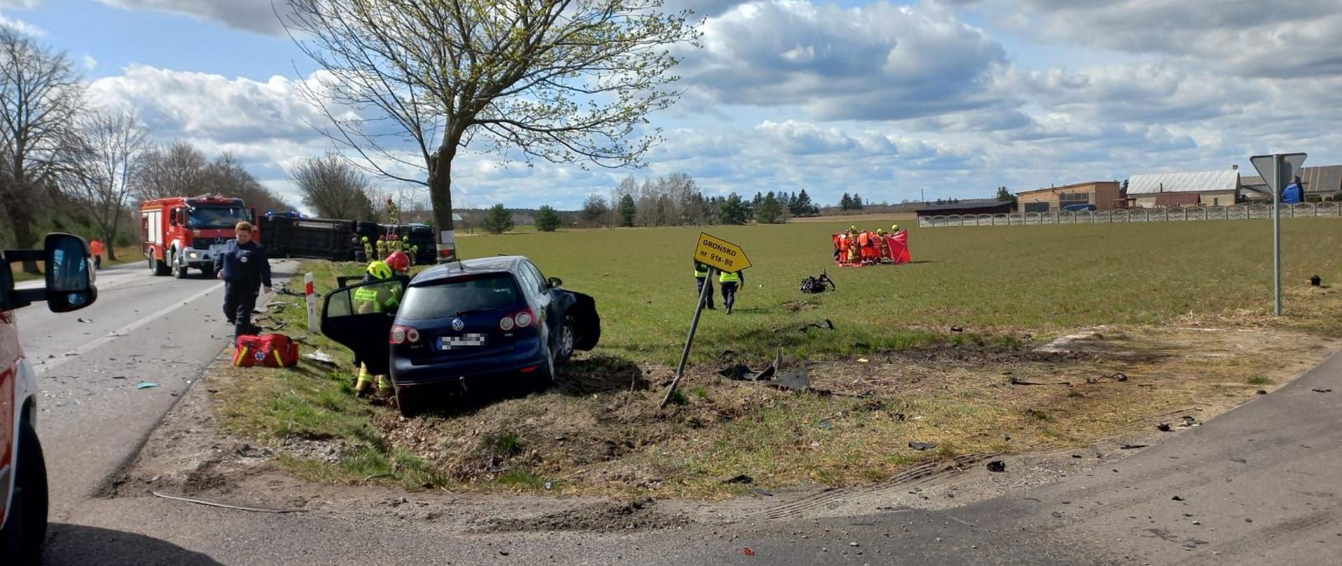 Na pierwszym palnie strażacy i policjanci prowadzący działania przy uszkodzonym samochodzie osobowym. Na drugim planie strażacy udzielający pomocy poszkodowanemu na przydrożnym polu oraz działający przy ciężarówce.
