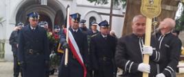 Pogrzeb strażaka. Kościół parafialny w Łaszczynie. Z kościoła na cmentarz zmierza kondukt pogrzebowy. Na pierwszym planie mężczyzna niosący krzyż, za nim poczet sztandarowy wystawiony przez KP PSP w Rawiczu oraz delegacje z wiązankami kwiatów.