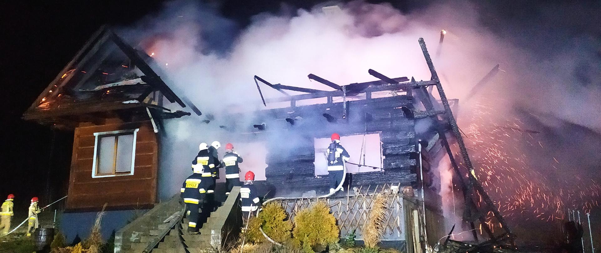 Zdjęcie przedstawia budynek objęty w całości ogniem. Wokół domu
widnieje żółta poświata. Na tle płomieni można zauważyć resztki więźby
dachowej. Widać również strażaków gaszących pożar.
