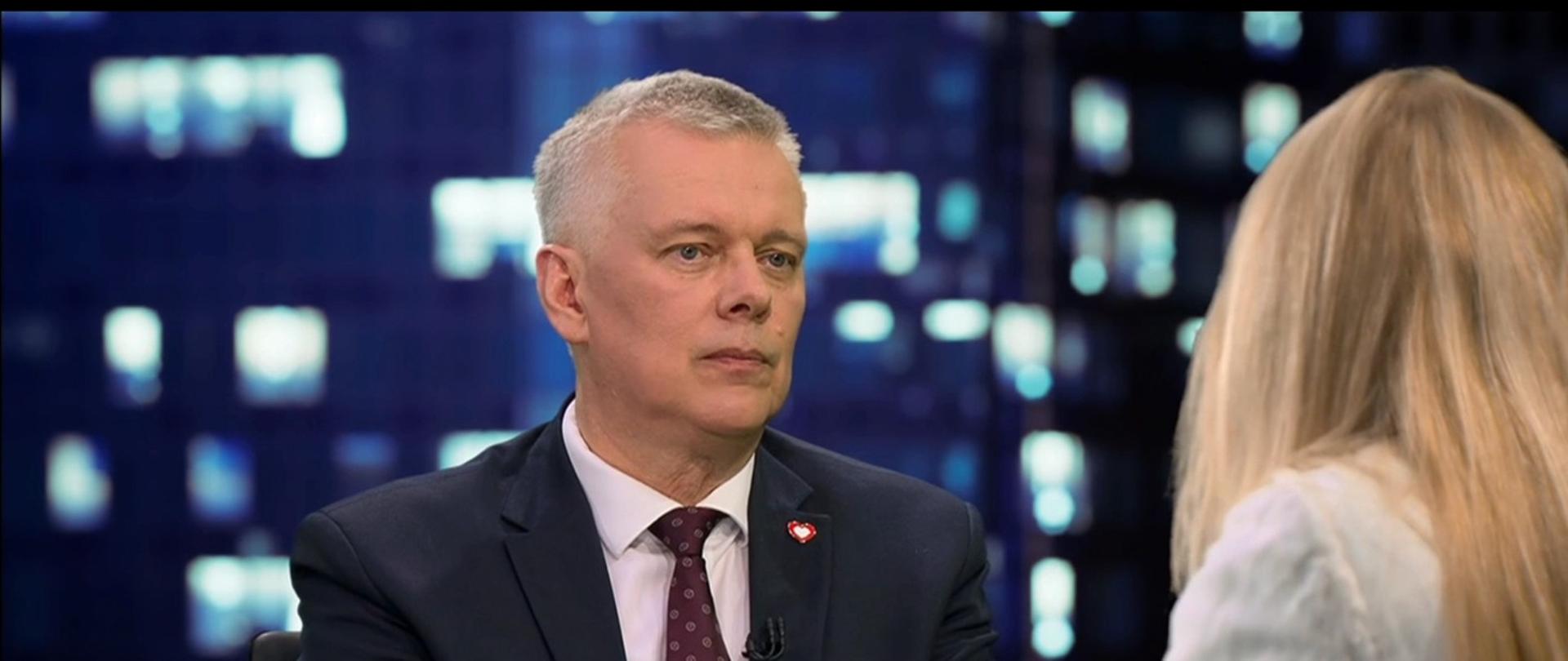 Kadr z programu informacyjnego TVN24 przedstawiający siedzącego w studiu ministra Tomasza Siemoniaka w granatowym garniturze, białej koszuli i bordowym krawacie. Rozmawia z panią Moniką Olejnik widoczną tyłem po prawej stronie kadru. W tle rozmyte światła studia telewizyjnego. Na dole ekranu widoczne są paski informacyjne TVN24 oraz napis „Wstajesz i Wiesz 7:10” i „Konrad Piasecki, TVN24"