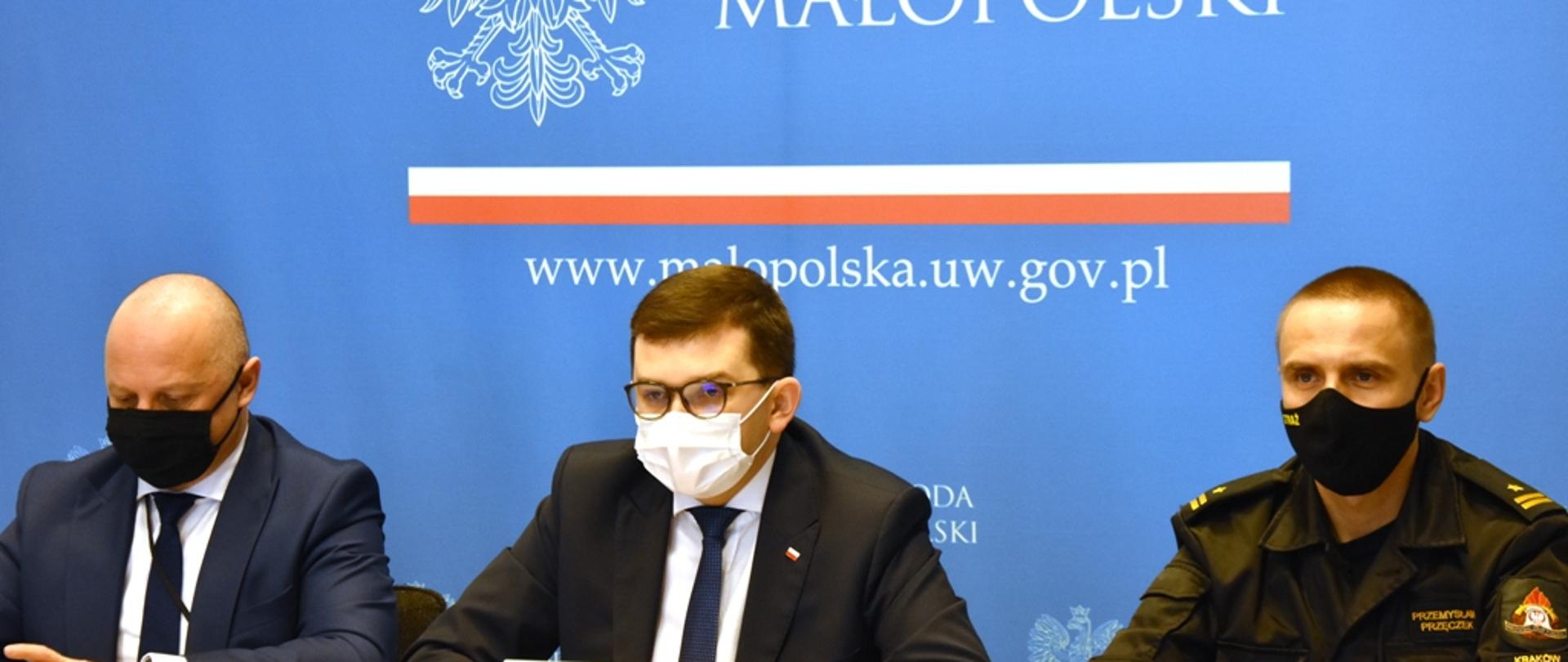 Przy stole siedzi trzech mężczyzn- m.in. Wojewoda Małopolski, Wszyscy panowie maja założone maseczki. Z-ca MKW PSP za nimi baner wojewody małopolskiego na niebieskim tle.