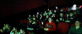 Młodzież siedzi w zaciemnionej sali kinowej na czerwonych fotelach, trzymając przekąski i napoje, oglądając film na ekranie. Uczniowie z internatu podczas seansu filmowego.