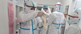 Na zdjęciu w pomieszczeniu z białymi ścianami i białym regałem po lewej stronie, widać pięć osób ustawionych przodem, bokiem i plecami, ubranych w białe kombinezony ochronne z zasłoniętymi białymi maseczkami twarzami i przeźroczystymi przyłbicami. Osoba na pierwszym planie zakłada białe rękawiczki ochronne, w tle osoba poprawiająca czepek na głowie.