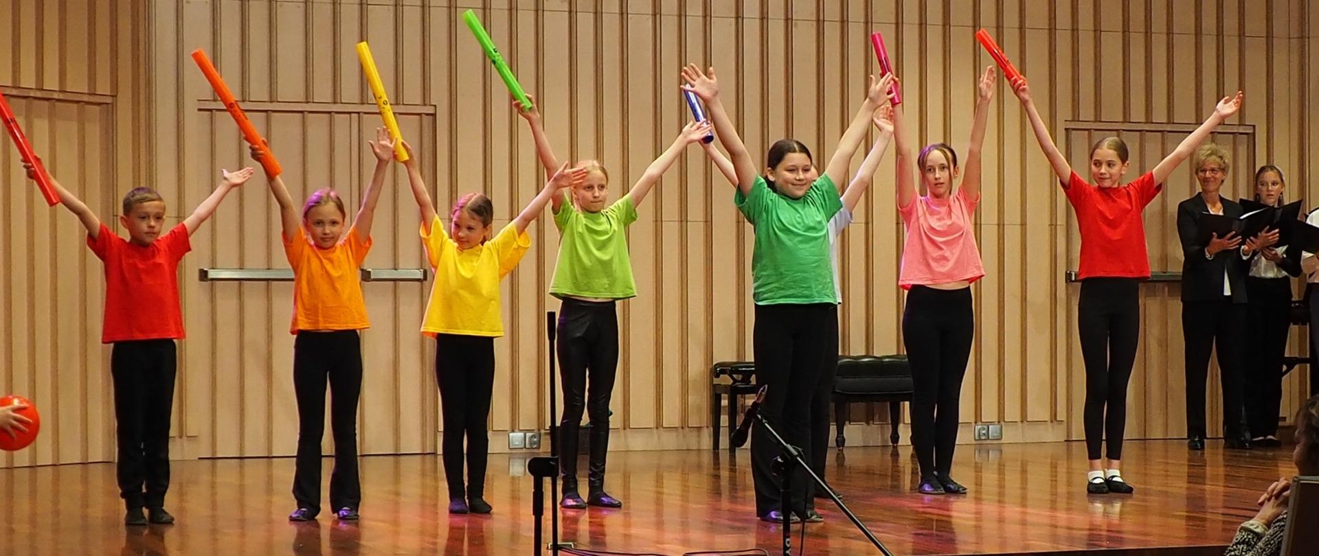 Dziecięca grupa rytmiczna z uniesionymi rękami w kolorowych strojach stoi na scenie w sali koncertowej.