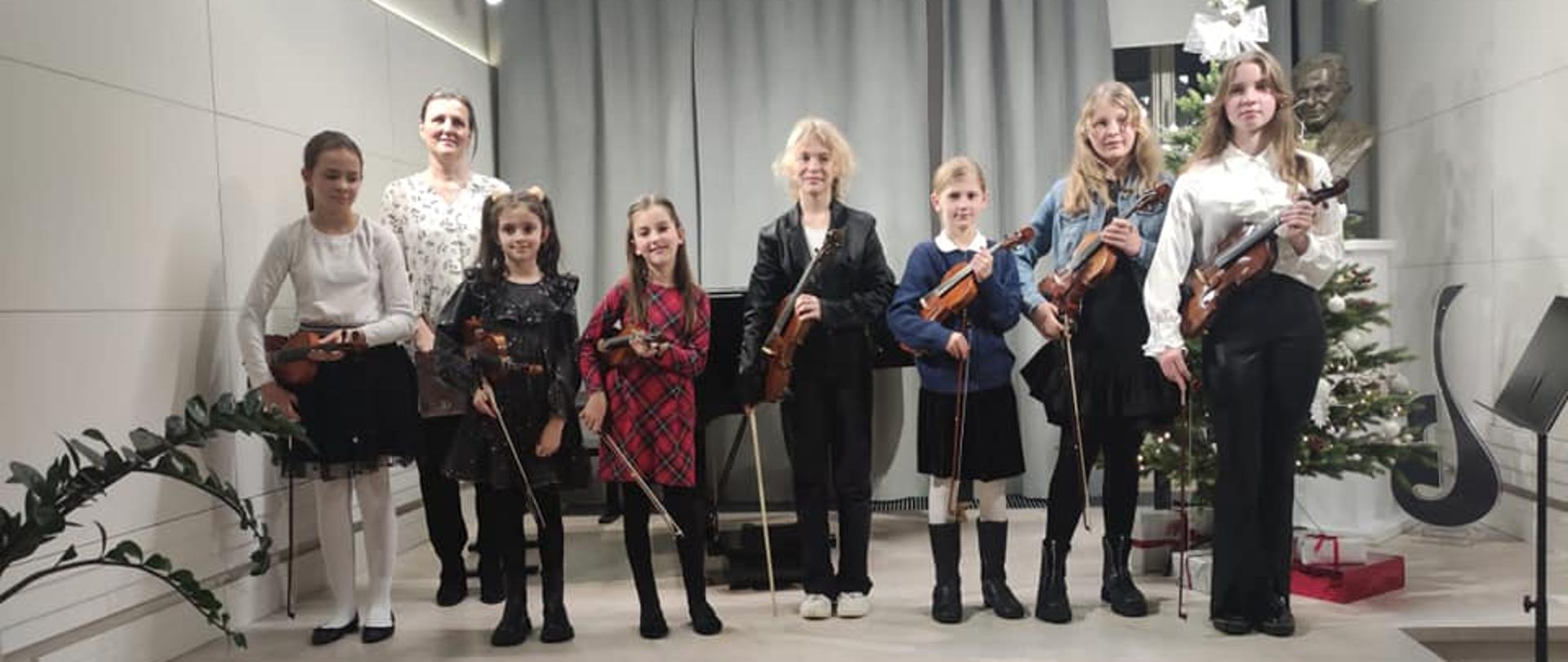 Zdjęcie przedstawia uczniów trzymających pod pachą skrzypce i stojących na scenie w jednym rzędzie. Od lewej za uczniami stoi akompaniator. W tle widać częściowo zasłonięte kotary w szarym kolorze. Z prawej strony za uczniami stoi ubrana choinka, a tuż obok niej postument z popiersiem patrona szkoły, statuetka logo szkoły i nieco z przodu pulpit czarny na nuty.