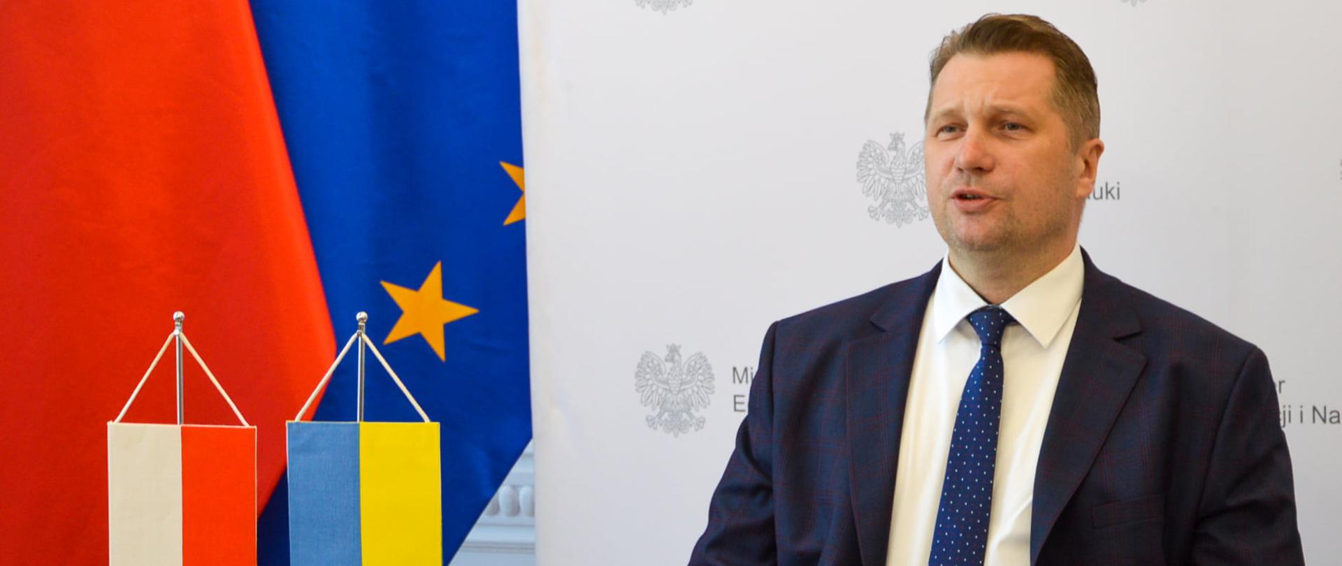 Minister Przemysław Czarnek siedzi przy biurku podczas wideokonferencji. Przed nim stoją flagi Polski i Ukrainy