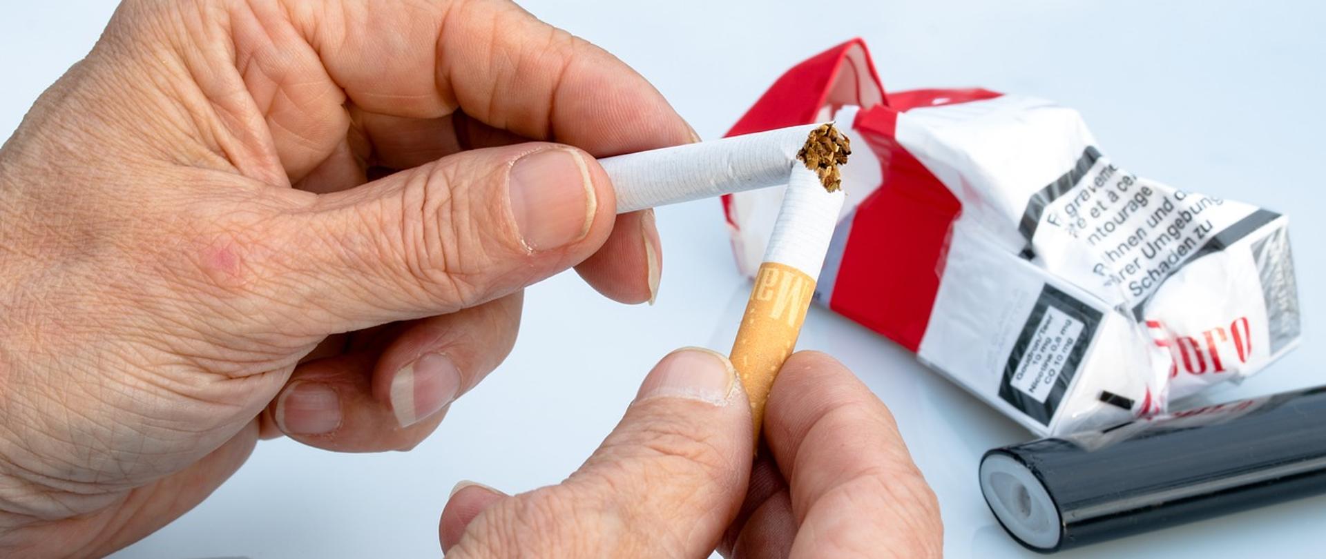 Zdjęcie przedstawia męskie ręce łamiące papierosa. W tle leży zapalniczka i zgnieciona paczka papierosów