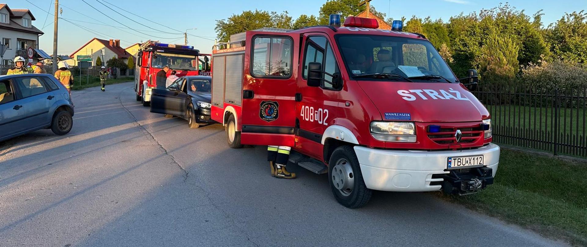 Samochody pożarnicze po prawej stronie, po lewej stronie niebieski samochód biorący udział w wypadku oraz strażacy. W tle domy