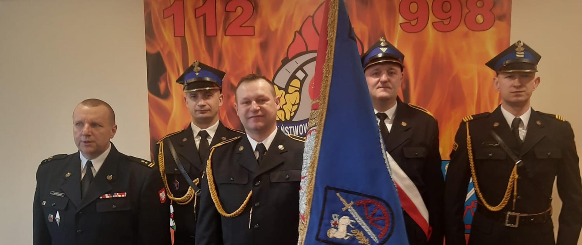 Zdjęcie zrobione na świetlicy. Na zdjęciu z lewej komendant powiatowy z pocztem sztandarowym i strażakiem, który żegna się ze służbą. Wszyscy w mundurach galowych.