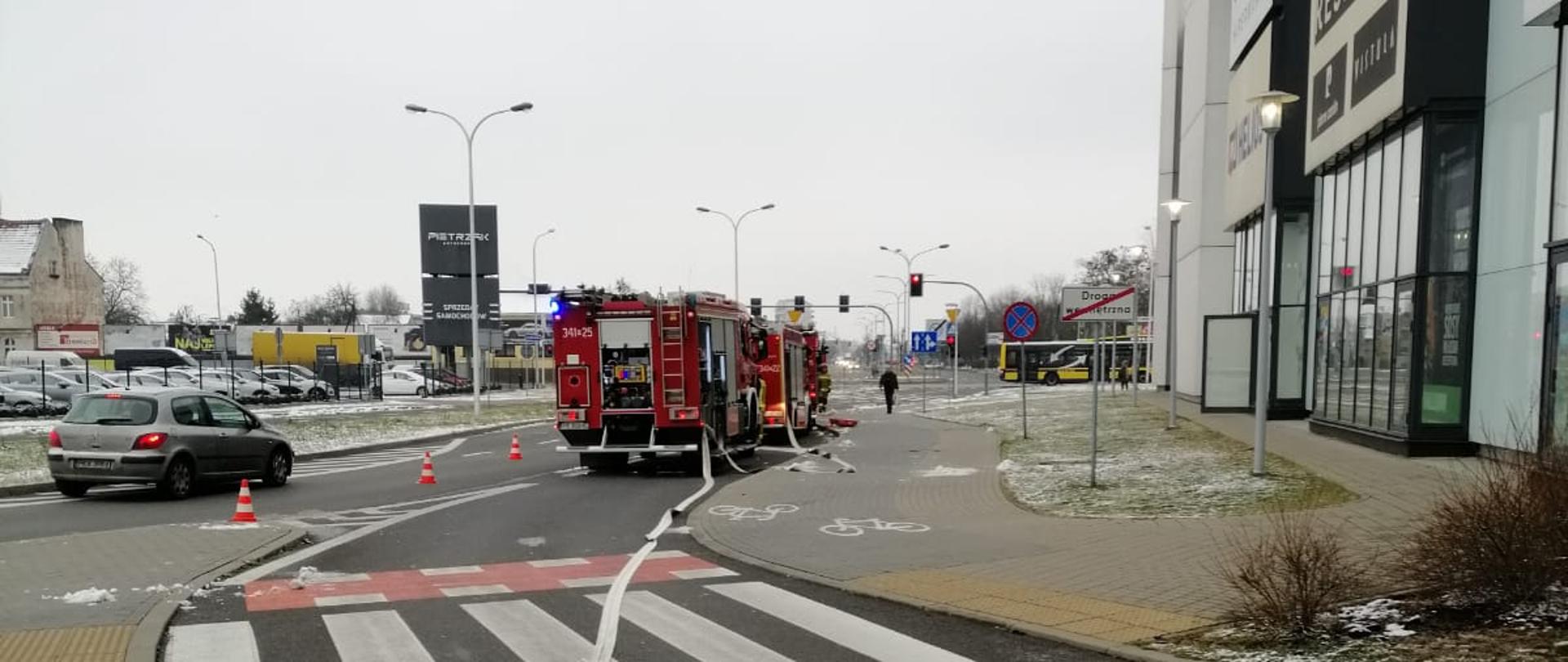 Na zdjęciu widać samochody gaśnicze w kolorze czerwonym, posiadają sygnalizację ostrzegawczą oraz numery operacyjne. Do samochodów podłączone są linie wężowe. Widać ścianę galerii po prawej stronie zdjęcia z reklamami. Linia prowadzona jest po czarnej, asfaltowej drodze. Zachmurzenie pełne. Na pierwszym planie przejście drogowe.