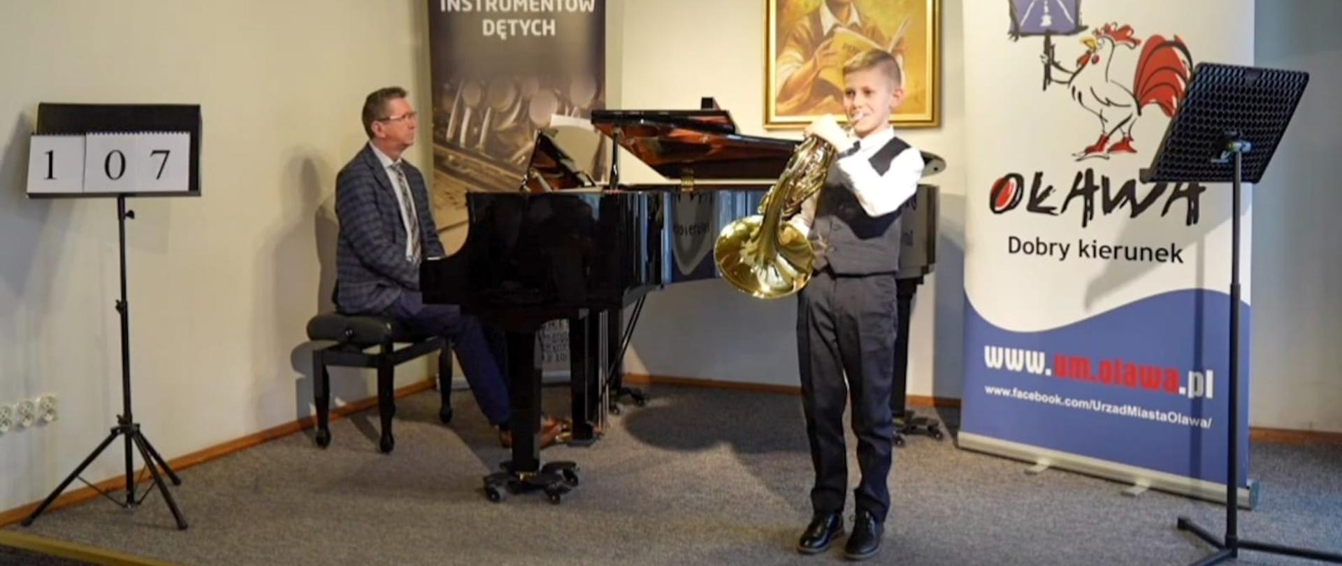 Uczeń gra na waltorni stojąc przed fortepianem, mężczyzna gra na fortepianie, w tle banery Konfrontacje Instrumentów Dętych oraz Urząd Miejski w Oławie - Oława dobry kierunek