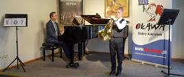 Uczeń gra na waltorni stojąc przed fortepianem, mężczyzna gra na fortepianie, w tle banery Konfrontacje Instrumentów Dętych oraz Urząd Miejski w Oławie - Oława dobry kierunek