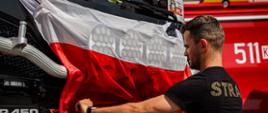Strażak zawiesza flagę Polski na masce samochodu ratowniczo - gaśniczego