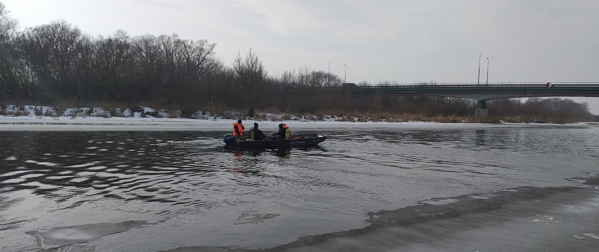 Na zdjęciu widoczna jest oblodzona z dwóch stron brzegów rzeka Narew, środkiem płynie woda. Strażacy w łodzi na środku rzeki płyną w miejsce w którym skoczył z mostu mężczyzna. Na łodzi widać dwóch nurków ubranych w skafandry nurkowe oraz sprzęt. Dwóch strażaków -ratowników ubranych jest w kamizelki ratunkowe. Ratownicy płyną pod most w miejscowości Piątnica.