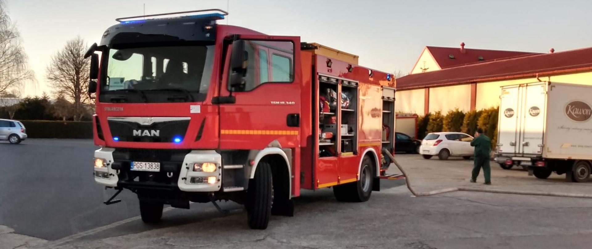 Zdjęcie przedstawia wóz strażacki podczas ćwiczeń. Samochód jest czerwony. Z samochodu podawana jest woda do działań gaśniczych.