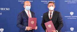 Na zdjęciu znajduje się dwóch, ubranych w garnitury mężczyzn. Mężczyźni Ci pozują na tle granatowej ścianki wizerunkowej Wojewody Mazowieckiego, prezentując w rękach brązowe teczki, w których znajdują się podpisane umowy. 