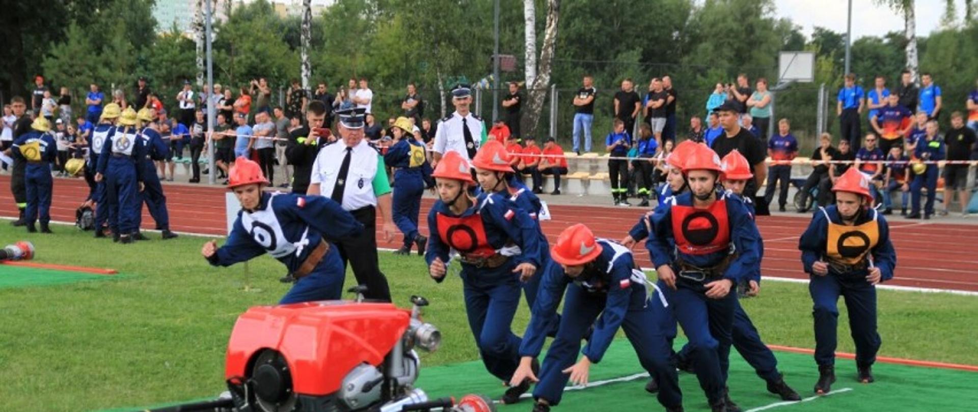 grupa ludzi w niebieskich kombinezonach i czerwnoych hełmach biegnie do sprzętu pożarniczego - pompa i węże , który stoi na murawie bosika, w tle trybuny z widzami 