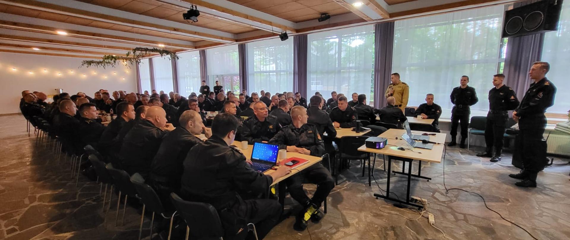 Na zdjęciu funkcjonariusze PSP siedzą przy stole na odprawie szkoleniowej. Na rzutniku wyświetlana jest prezentacja. Przemawia Z-ca Wielkopolskiego Komendanta Wojewódzkiego PSP..