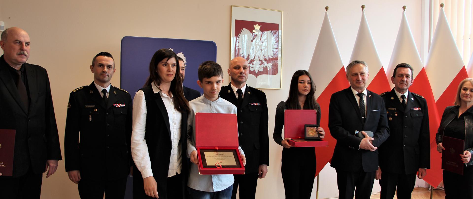 Pamiątkowe zdjęcie grupowe. Odznaczeni odznaką Iuvenis Forti: Zosia Cerkowniak i Patryk Opałko oraz wojewoda świętokrzyski Zbigniew Koniusz, zastępca świętokrzyskiego komendanta wojewódzkiego Państwowej Straży Pożarnej st. bryg. Grzegorz Rajca, komendant miejski PSP w Kielcach st. bryg. Sławomir Karwat, komendant powiatowy st. bryg. Artur Brachowicz, dyrektor II LO im. Jana Śniadeckiego w Kielcach Tomasz Kośny i inni.