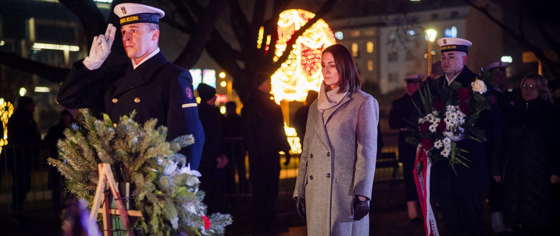 kobieta (wojewoda) i dwóch marynarzy niosą wieniec pod pomnik Poległych stoczniowców