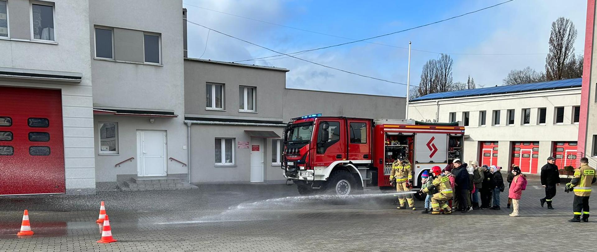 Na placu stoi samochód strażacki. Samochód ma kolor czerwony i czarny. Obok samochodu stoi grupka dzieci oraz funkcjonariusze PSP. Chłopiec leje wodę z węża strażackiego w kierunku trzech pachołków. Obok chłopca kucnął strażak w czerwonym hełmie. W tle widać budynek komendy straży pożarnej. Po lewej stronie widać kawałek czerwonej bramy garażowej. Okna
i białe drzwi. Po prawej stronie widać budynek straży pożarnej, okna i czerwone bramy garażowe. Po prawej stronie za budynkiem widać drzewa.
