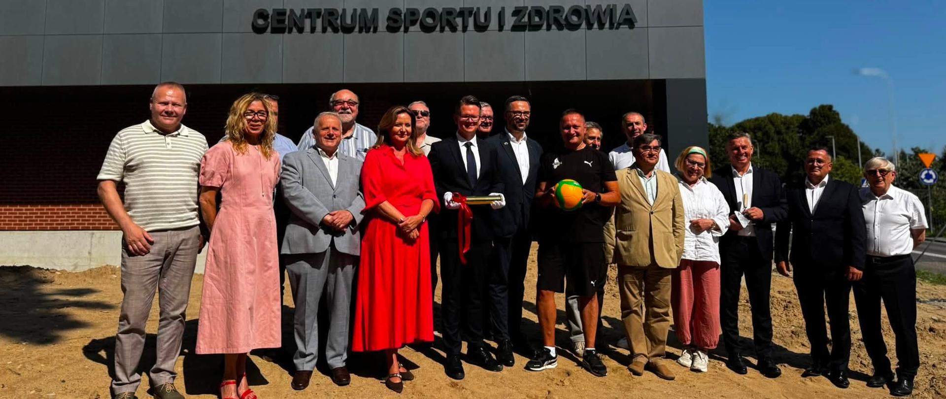 Grupa oficjeli uczestniczących w wydarzeniu pozuje do wspólnego zdjęcia na tle Wielkiego Centrum Sportu i Zdrowia. Wśród uczestników znajdują się przedstawiciele władz oraz zaproszeni goście.