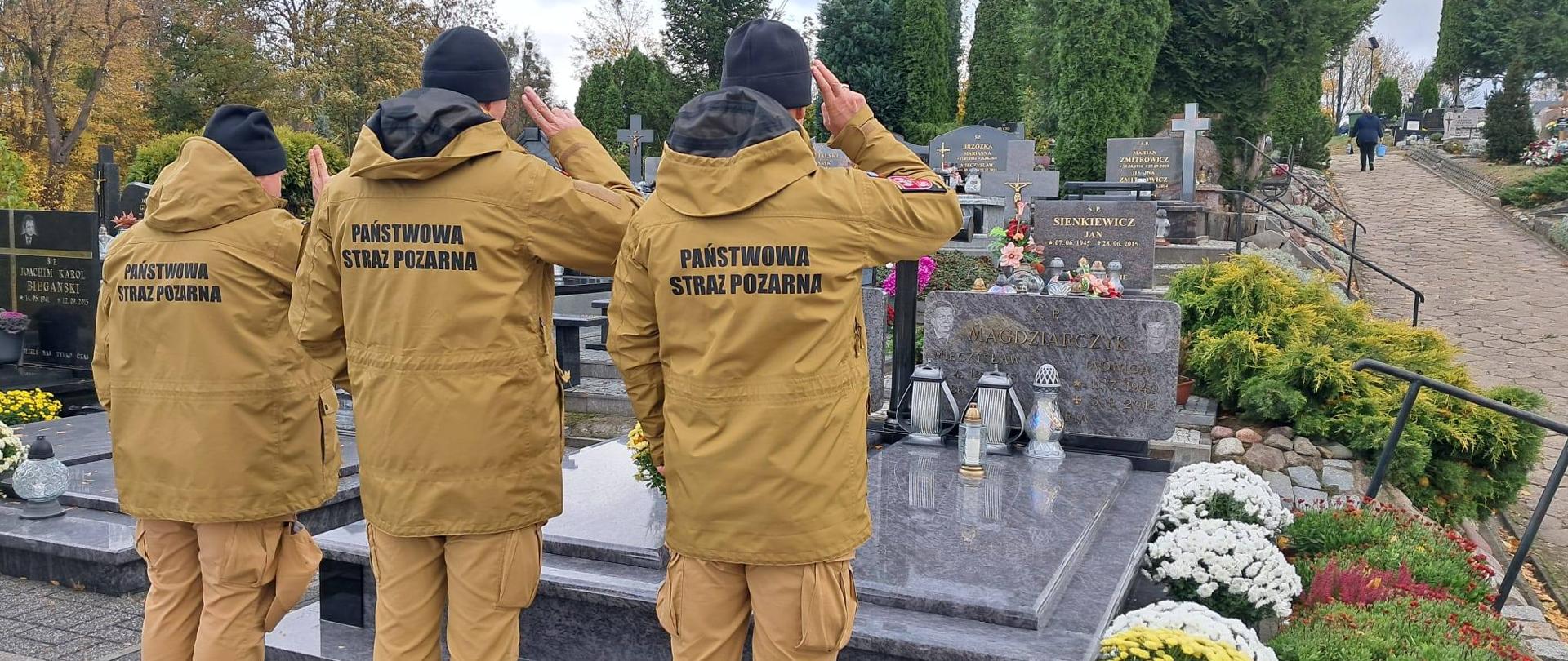 Trzech strażaków w jasnych mundurach i czarnych czapkach stoją przed grobem na cmentarzu.