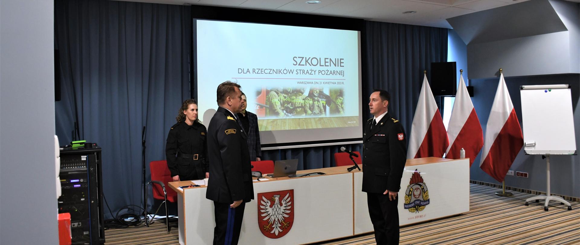 Naczelnik Wydziału Organizacji i Nadzoru Komendy Wojewódzkiej PSP w Warszawie składa meldunek Mazowieckiemu Komendantowi Wojewódzkiemu PSP. Znajdują się na sali konferencyjnej. W tle wyświetlona jest prezentacja na ekranie.