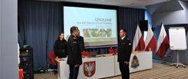 Naczelnik Wydziału Organizacji i Nadzoru Komendy Wojewódzkiej PSP w Warszawie składa meldunek Mazowieckiemu Komendantowi Wojewódzkiemu PSP. Znajdują się na sali konferencyjnej. W tle wyświetlona jest prezentacja na ekranie.