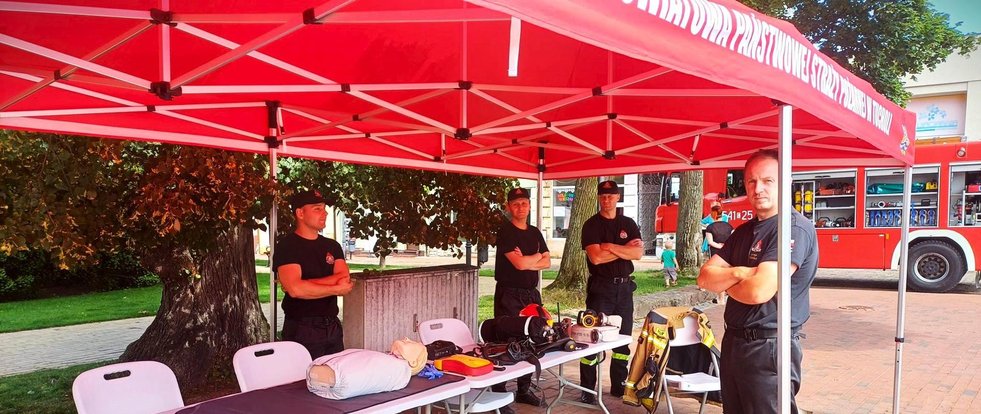 Czterech strażaków stoi pod czerwonym namiotem. Strażacy mają na sobie czarny mundur. Trzech strażaków ma ubraną czarną czapkę z daszkiem i stoją za białym stolikiem i białymi krzesłami. Jeden strażak stoi za stolikiem obok szarego słupka namiotu. Na stoliku widać sprzęt strażacki. Za strażakami widać drzewo, chodnik i trawnik z krzewami. Z prawej strony za strażakami widać czerwony wóz strażacki. Strażacy stoją na placu. 