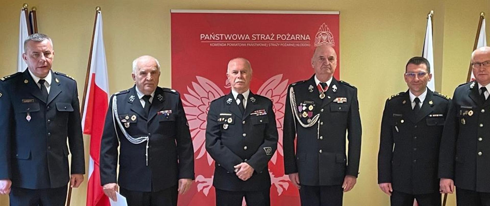 Sala Konferencyjna. W szeregu stoją druhowie Ochotniczej Straży Pożarnej,Komendant Powiatowy PSP w Kłodzku oraz Zastępca Komendanta Powiatowego PSP w Kłodzku. Za nimi baner z napisem "Państwowa Straż Pożarna" oraz godłem Polski.