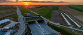 Widok z lotu ptaka na wlot do tunelu drogowego otoczony polami uprawnymi; nad horyzontem zachodzi słońce, oświetlając drogę i krajobraz ciepłym, pomarańczowym światłem.