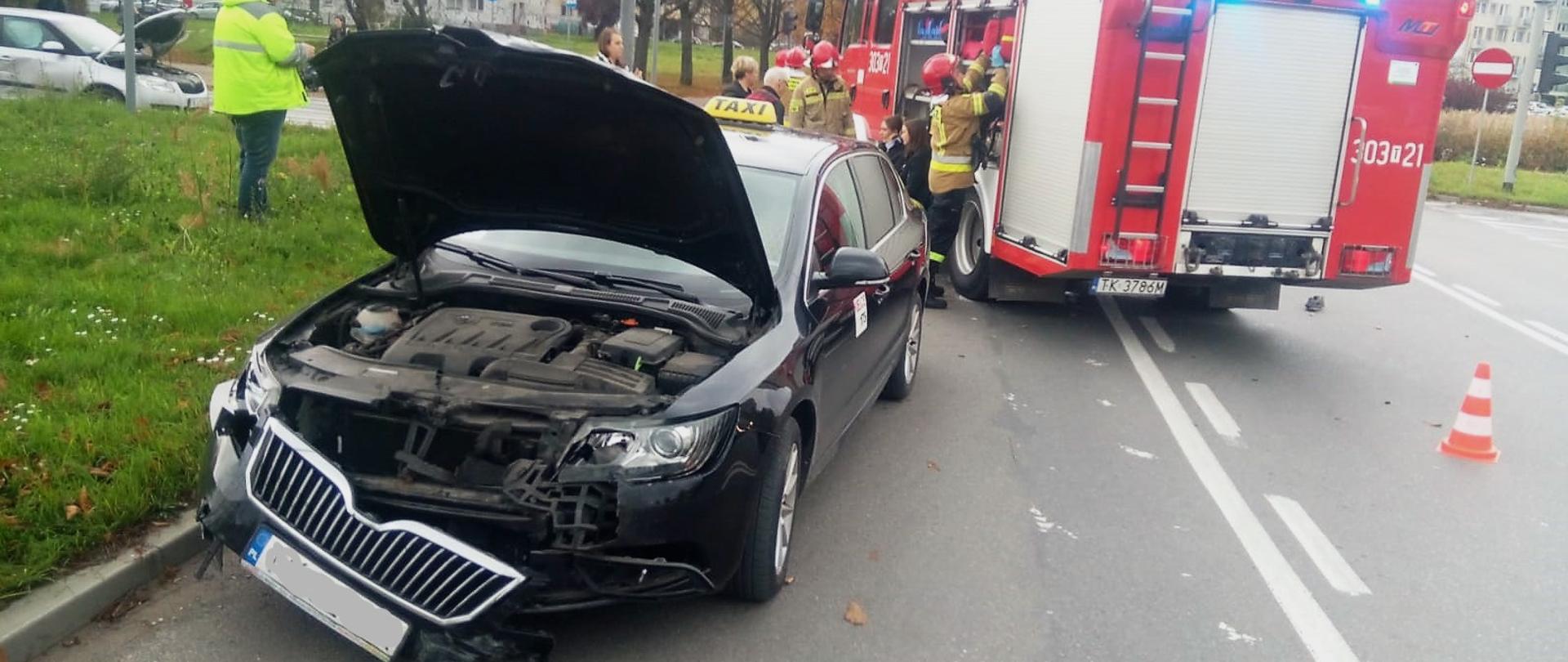 Zdjęcie przedstawia na pierwszym planie jeden z samochodów, które brały udział w zderzeniu. Z prawej strony stoi wóz pożarniczy zaś z lewej głęboko w tle drugi z uszkodzonych aut.