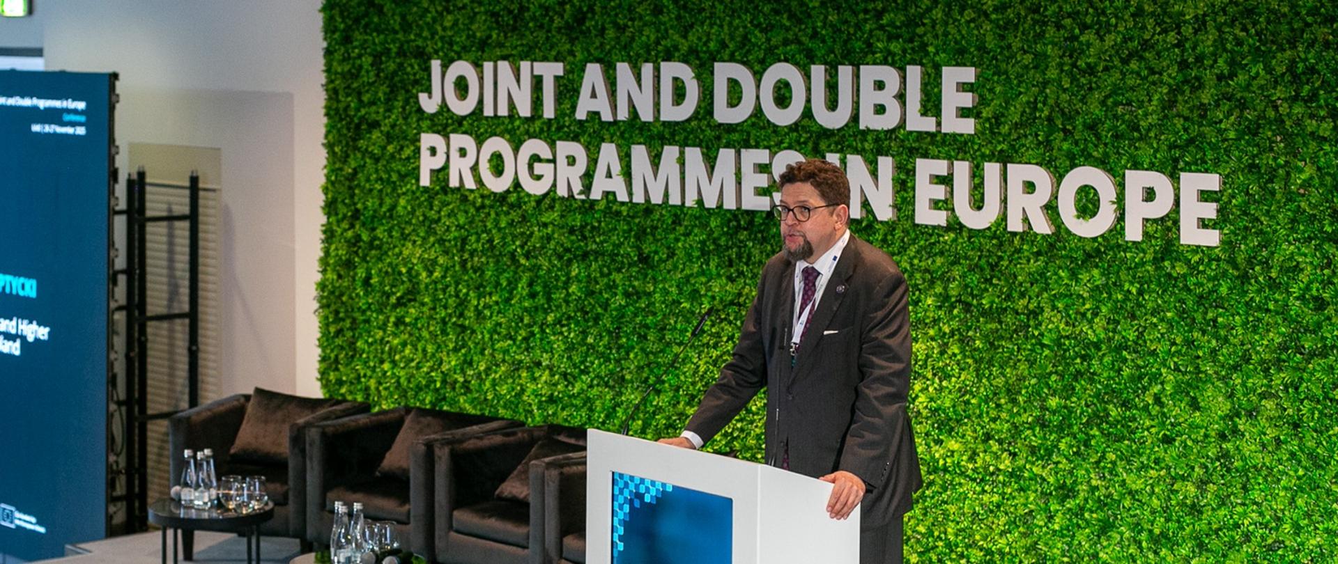 Zdjęcie z boku pod kątem, za mównicą stoi wiceminister Szeptycki i mówi do mikrofonu, za nim zielona ściana z białym napisem "Joint and double programme in Europe".