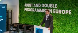 Zdjęcie z boku pod kątem, za mównicą stoi wiceminister Szeptycki i mówi do mikrofonu, za nim zielona ściana z białym napisem "Joint and double programme in Europe".