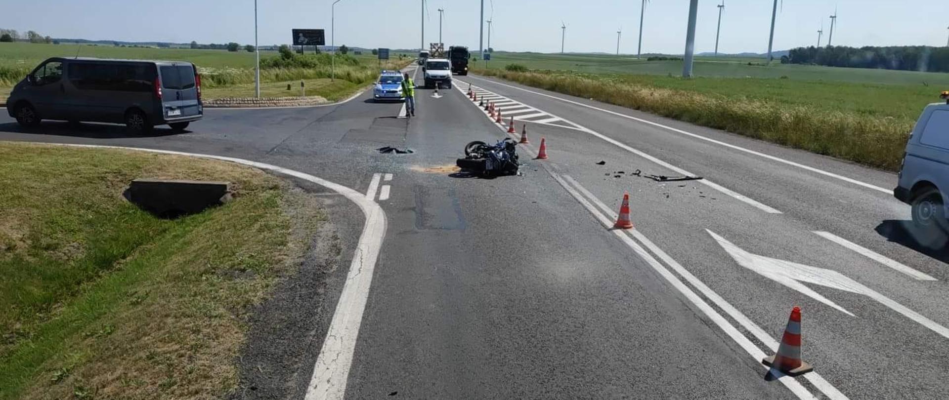 Dzień. Zjazd z drogi krajowej DK94 na stacje paliw. Na środku zjazdu rozbity motocykl. W głębi patrol policji blokuje ruch.