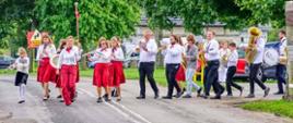 Ujęcie z bliska na przednią część orkiestry dętej występującej na trawiastym placu. Z lewej strony widoczna jest tamburmajorka w czerwonych spodniach dająca sygnał do marszu. Za nią idą młode dziewczęta i kobiety w czerwonych spódnicach, grające na fletach, klarnetach i talerzach, a w dalszych rzędach mężczyźni grający na trąbkach i saksofonach. W tle znajdują się namioty gastronomiczne oraz uczestnicy imprezy