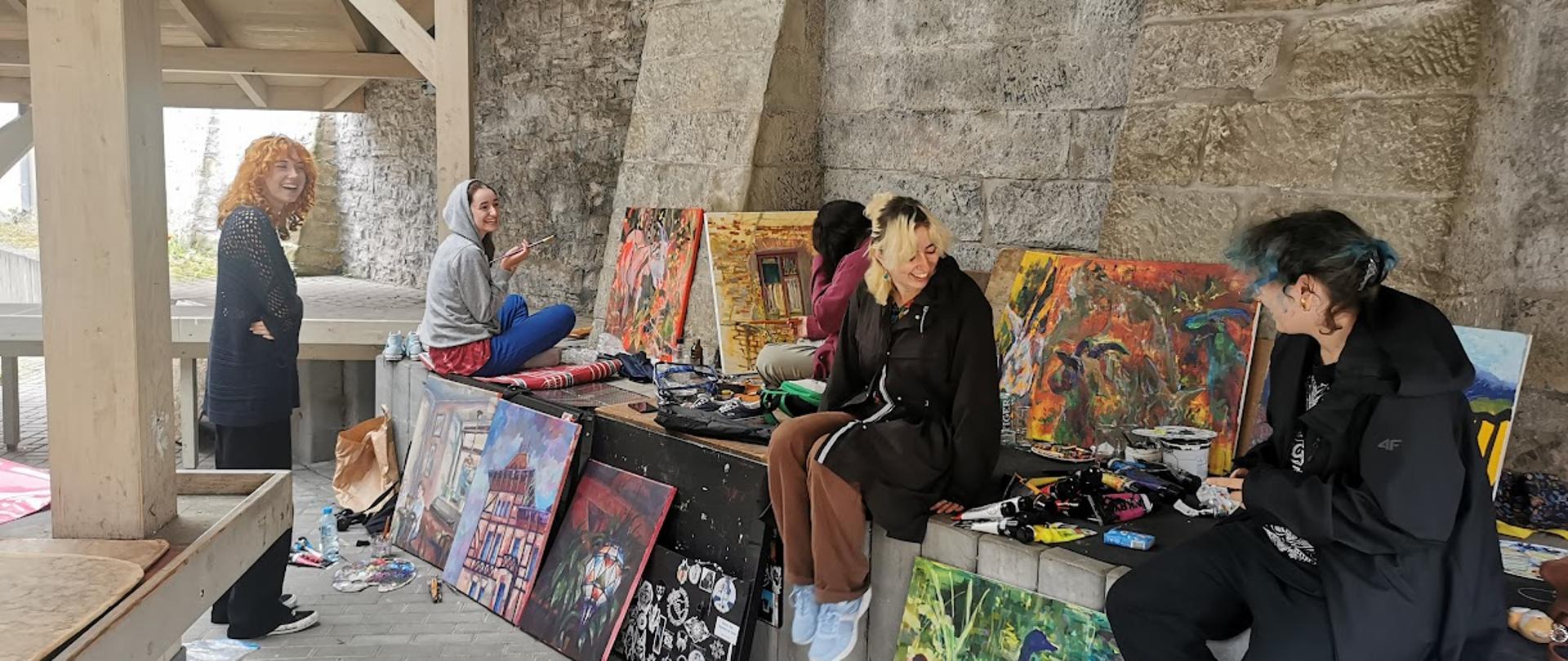 Pięć młodych dziewczyn uśmiechniętych stoją i siedzą między obrazami opartymi o mur