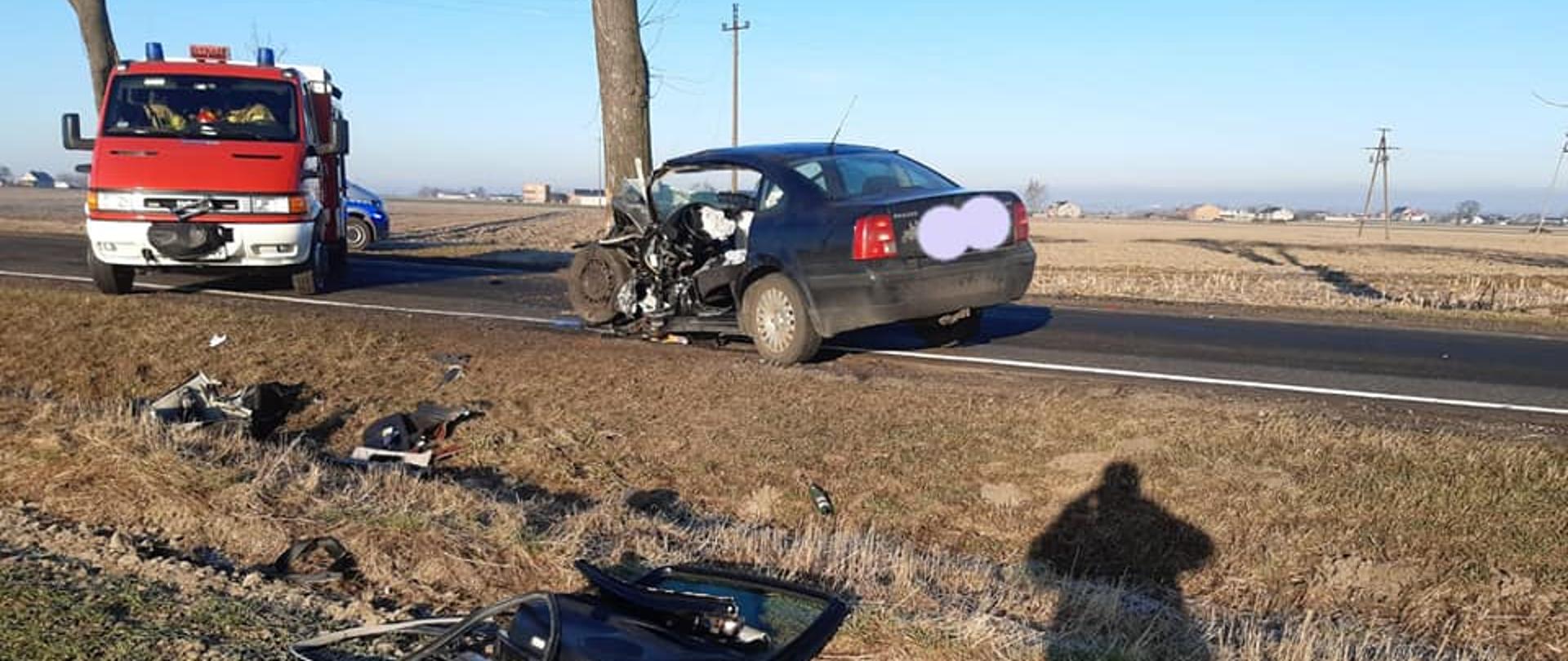 Na zdjęciu widać rozbity ciemny samochód osobowy, który stoi na jezdni. Obok niego stoi czerwony samochód strażacki. W rowie oraz na polu leżą wyrwane elementy samochodu. Za samochodem strażackim widać przód radiowozu. W tle widać drzewa oraz zabudowania. Na poboczu oraz rowie widać cień osoby robiącej zdjęcie. Jest widno.