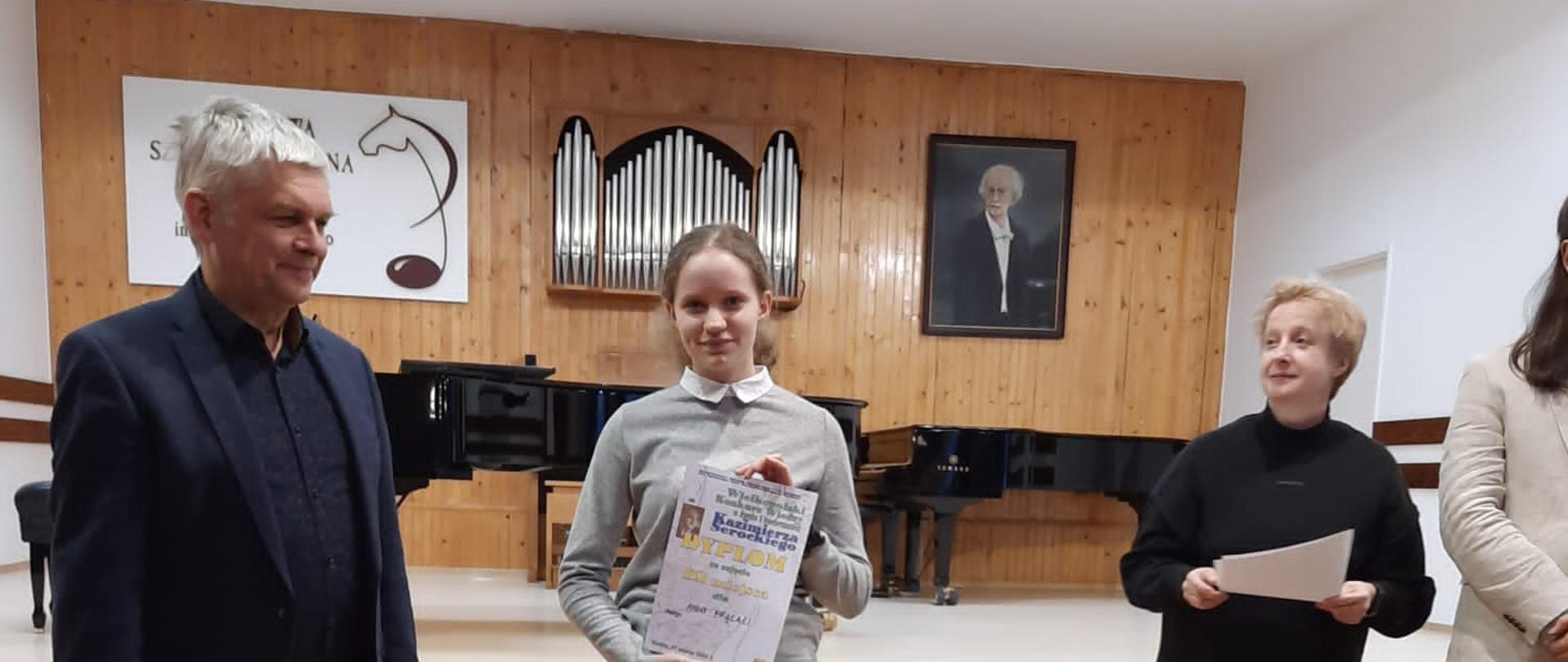 Laureatka III nagrody Wielkopolskiego Konkursu wiedzy o życiu i twórczości Kazimierza Serockiego w Koninie, obok dyrektor PSM w Koninie oraz przewodnicząca Jury, stoją na scenie sali koncertowej