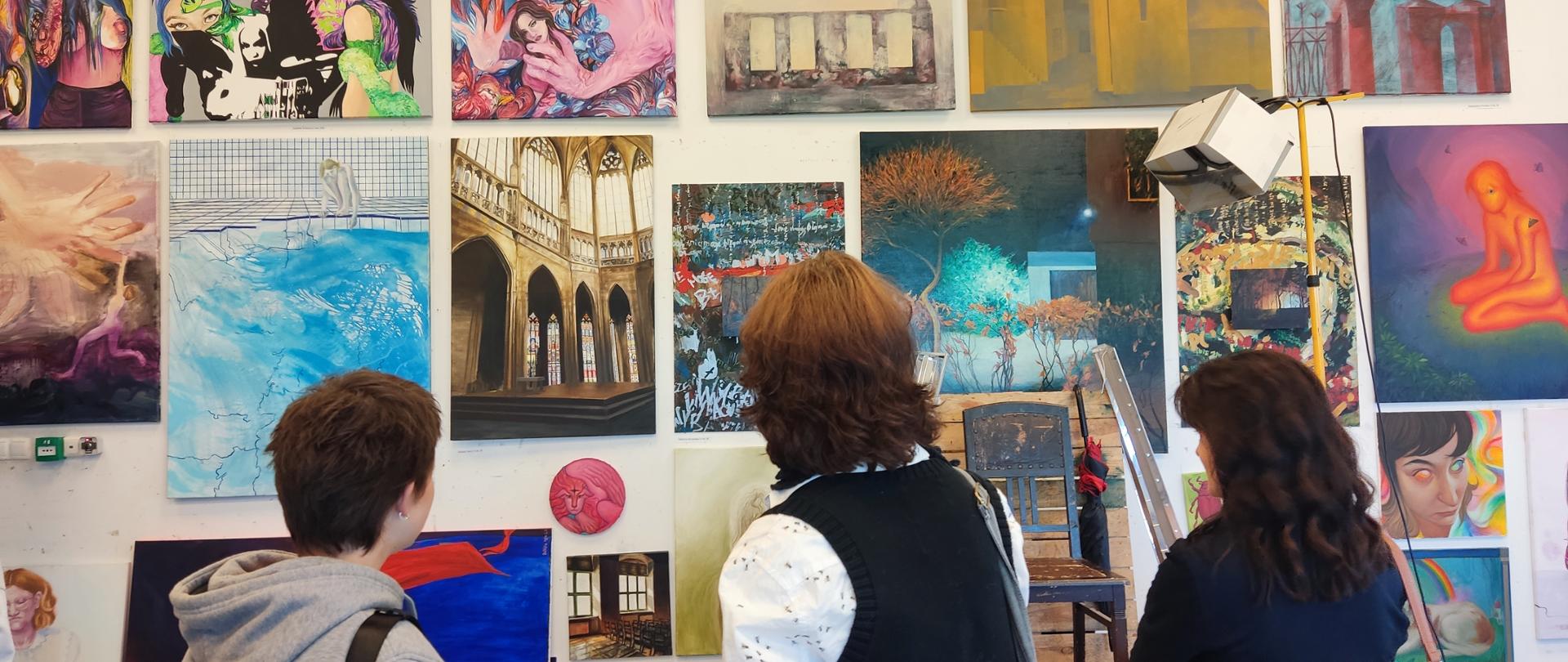 Trzy osoby stoją tyłem, patrzą na wystawę prac malarskich