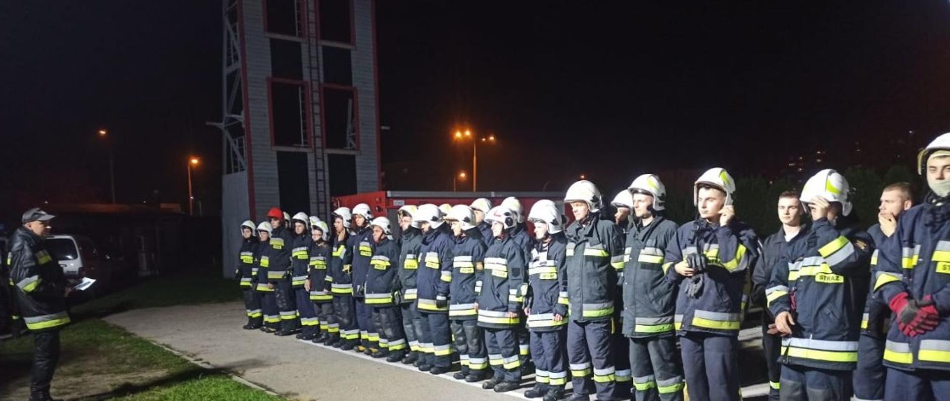 Na zdjęciu w porze nocnej przed siedzibą KP PSP w Ostrowcu Św. stoją na zbiórce przyszli ratownicy OSP - druhowie OSP. Przed nimi stoi oficer PSP, który tłumaczy zasady egzaminu praktycznego. W tle wspinalnia wykorzystywana do celów sportów pożarniczych.