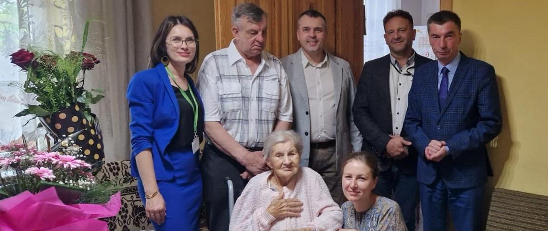 Starsza kobieta siedzi na wózku obok niej przykucnęła kobieta z tyłu stoi druga kobieta i czterech mężczyzn