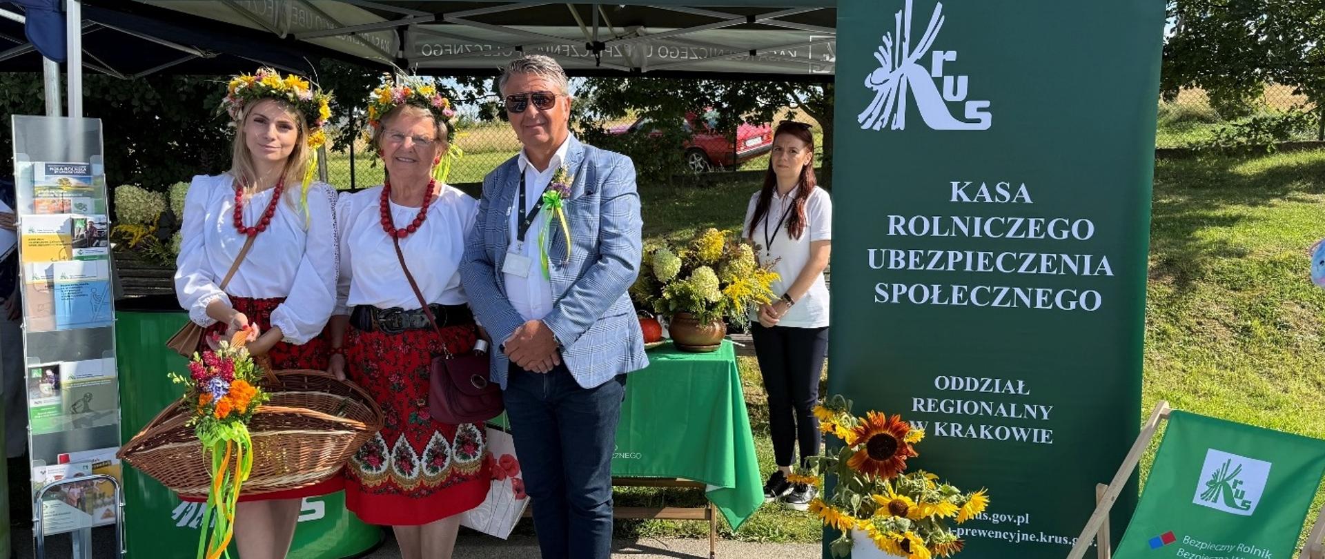 Stoisko informacyjno-prewencyjne podczas Dożynek Gminno-Parafialnych w Sułoszowej. Przed namiotem z logo KRUS znajdują się panie z KGW w strojach regionalnych, obok nich stoją pracownicy Placówki Terenowej KRU w Wolbromiu. Przed namiotem wystawiono bukiet ze słoneczników oraz leżak.